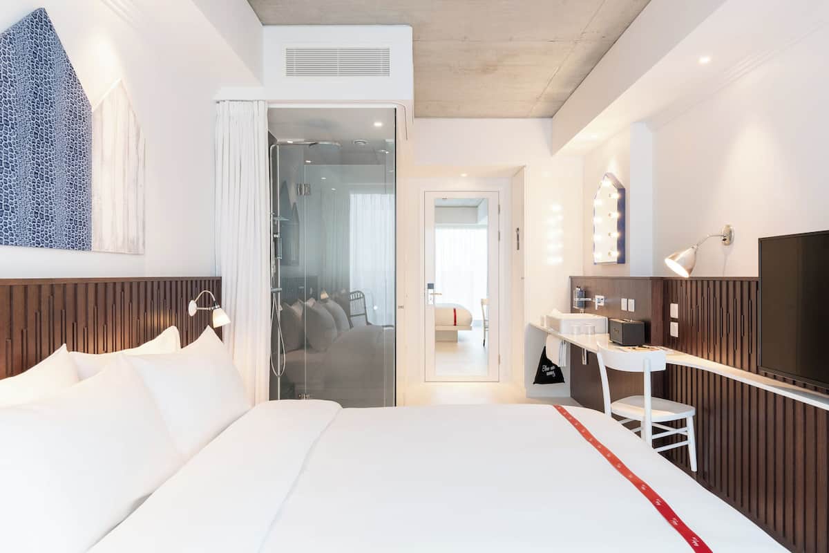 A hotel room with a bed, a vanity with a mirror, a television, and a shower area.