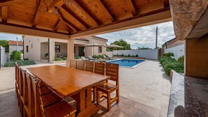 Une terrasse extérieure avec une table et des chaises en bois, une piscine et un espace abrité doté d’un plafond en bois.