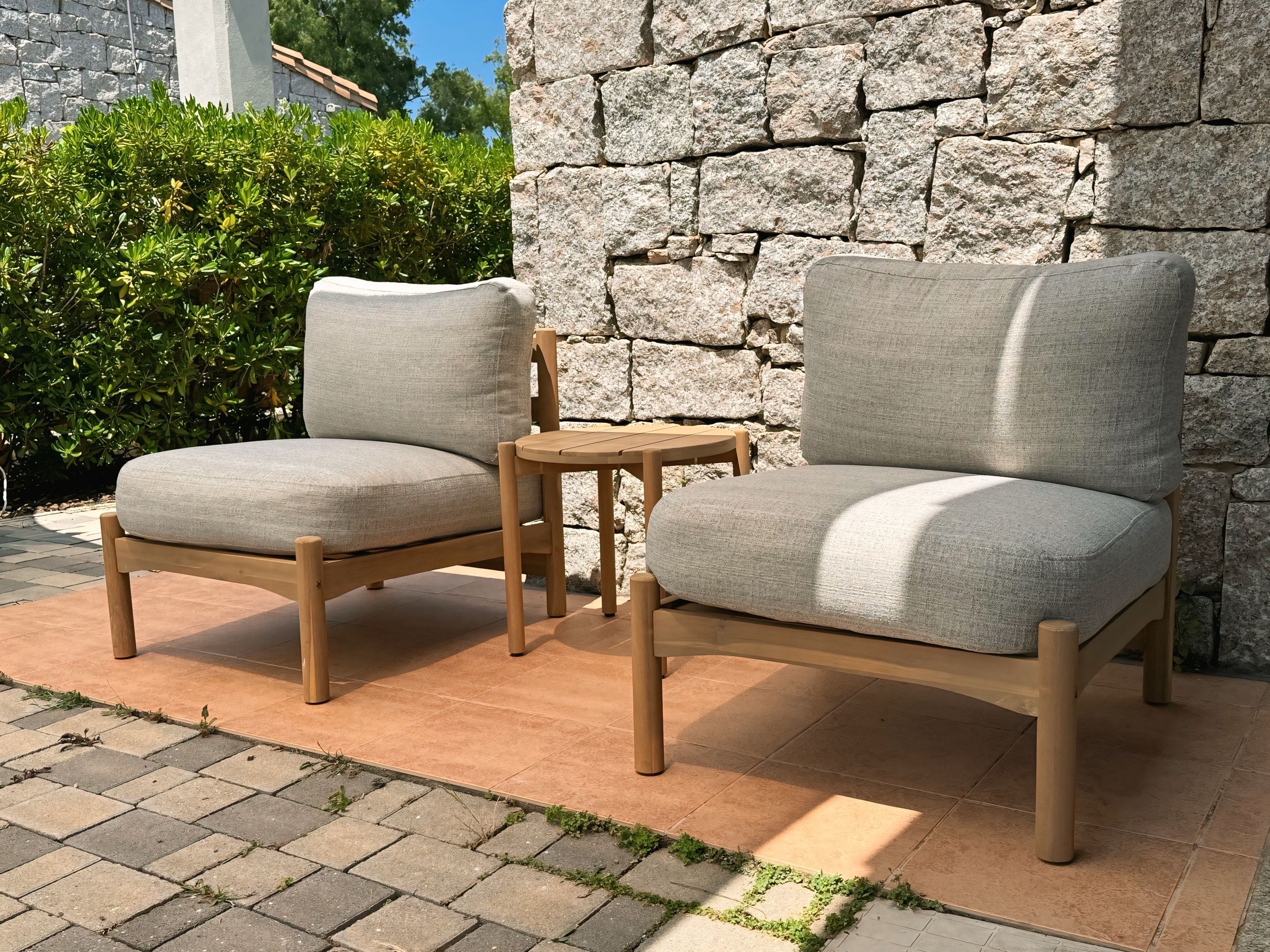 Dos sillones grises tapizados con armazones de madera y una pequeña mesa de madera, situados en un área pavimentada con un muro de piedra y vegetación de fondo.