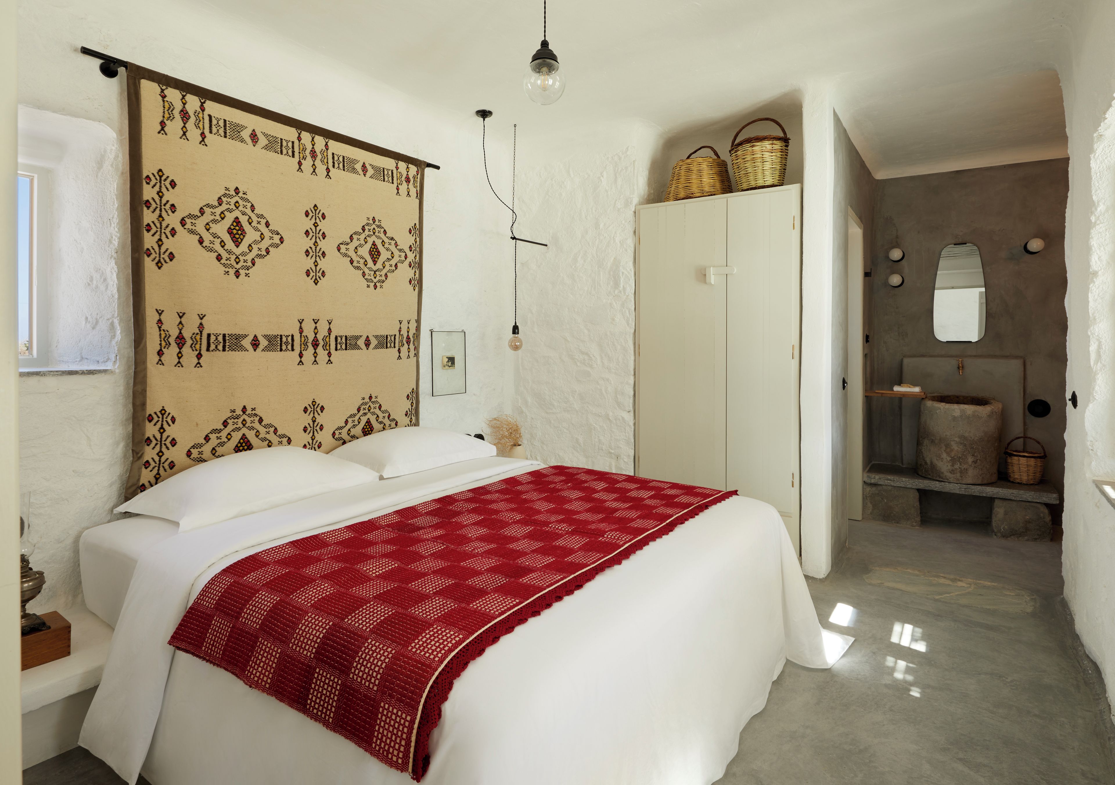 A bedroom with a bed, a wardrobe, a mirror, and a bathroom with a sink and a large container.