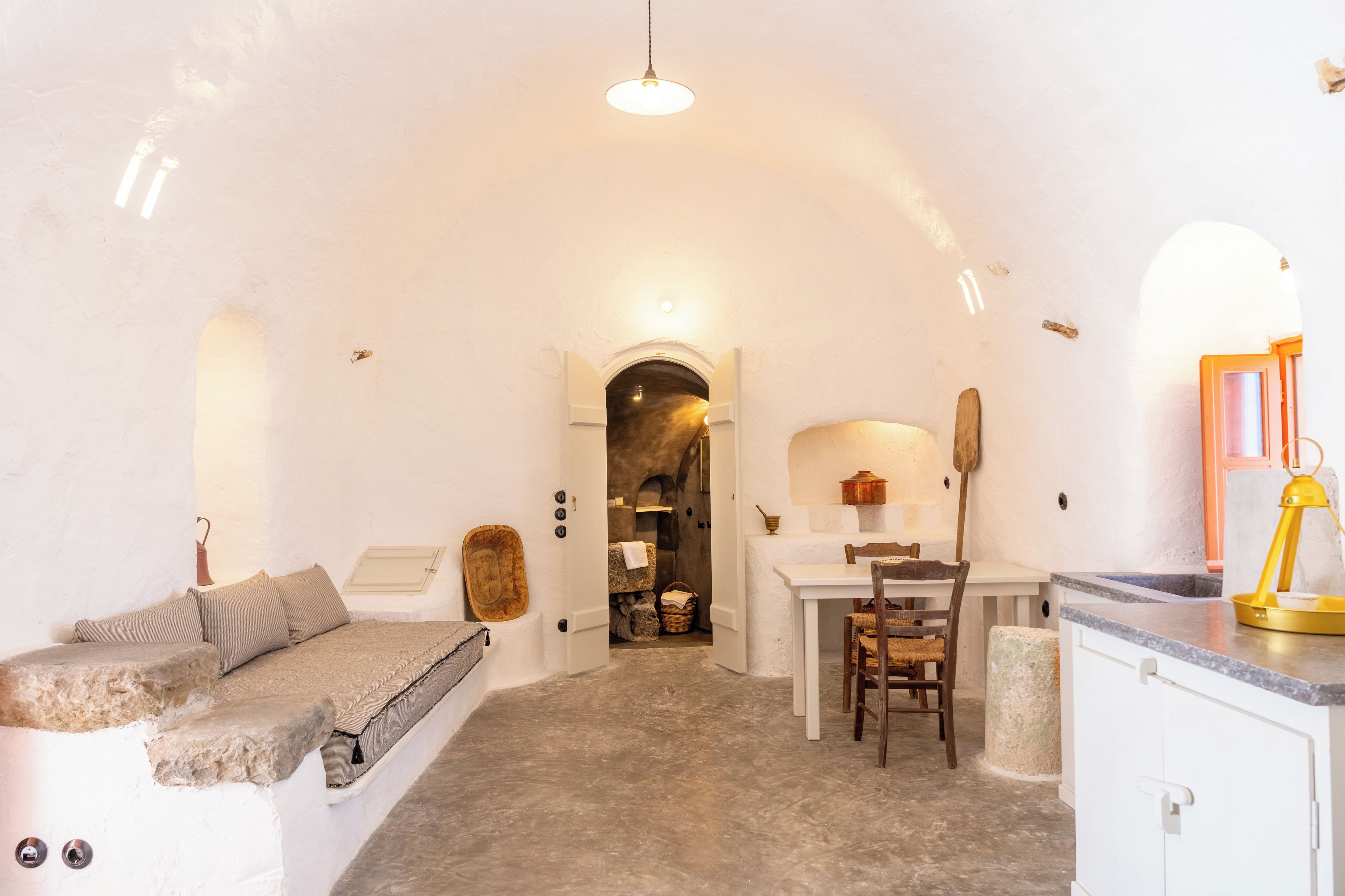 A cozy room with a stone bench, a small table, and a chair. There's a doorway leading to a kitchen area with a stove and a sink.