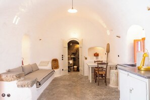 A cozy room with a stone bench, a small table, and a chair. There's a doorway leading to a kitchen area with a stove and a sink. - Santa Irini Retreat (Therasia)