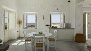 A room with a white table and chairs, a small wooden cabinet, and a view of the sea through open windows.