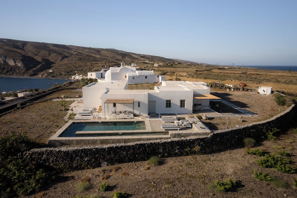 A white villa with a swimming pool, surrounded by a stone wall, overlooking a coastal landscape.