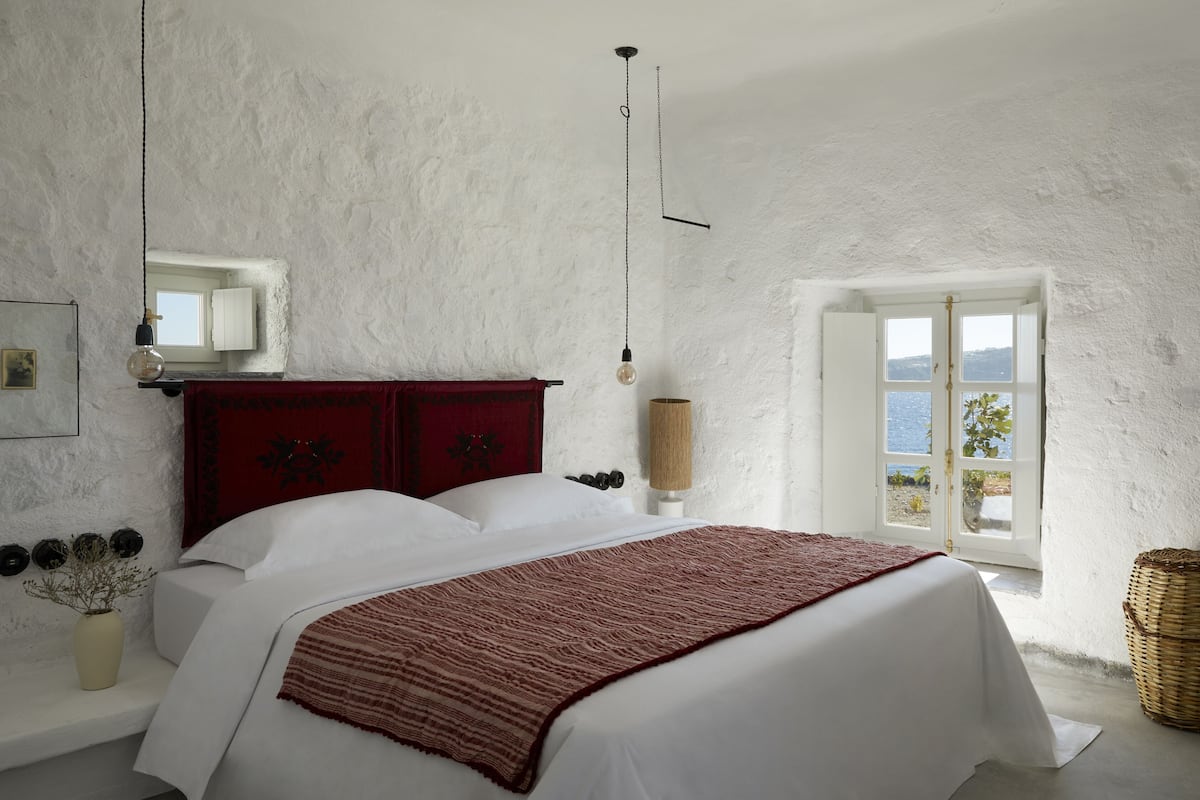 A bedroom with a bed, a red headboard, a bedside lamp, a window with a view, and a wall-mounted shelf with decorative items.