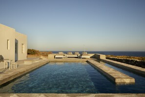 A rectangular pool with clear blue water, surrounded by stone walls and a white building with a door and windows. - Santa Irini Retreat (Therasia)