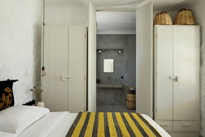 A bedroom with a bed, a wardrobe, a hanging light fixture, and a small bathroom with a mirror and sink. - Santa Irini Retreat (Therasia)