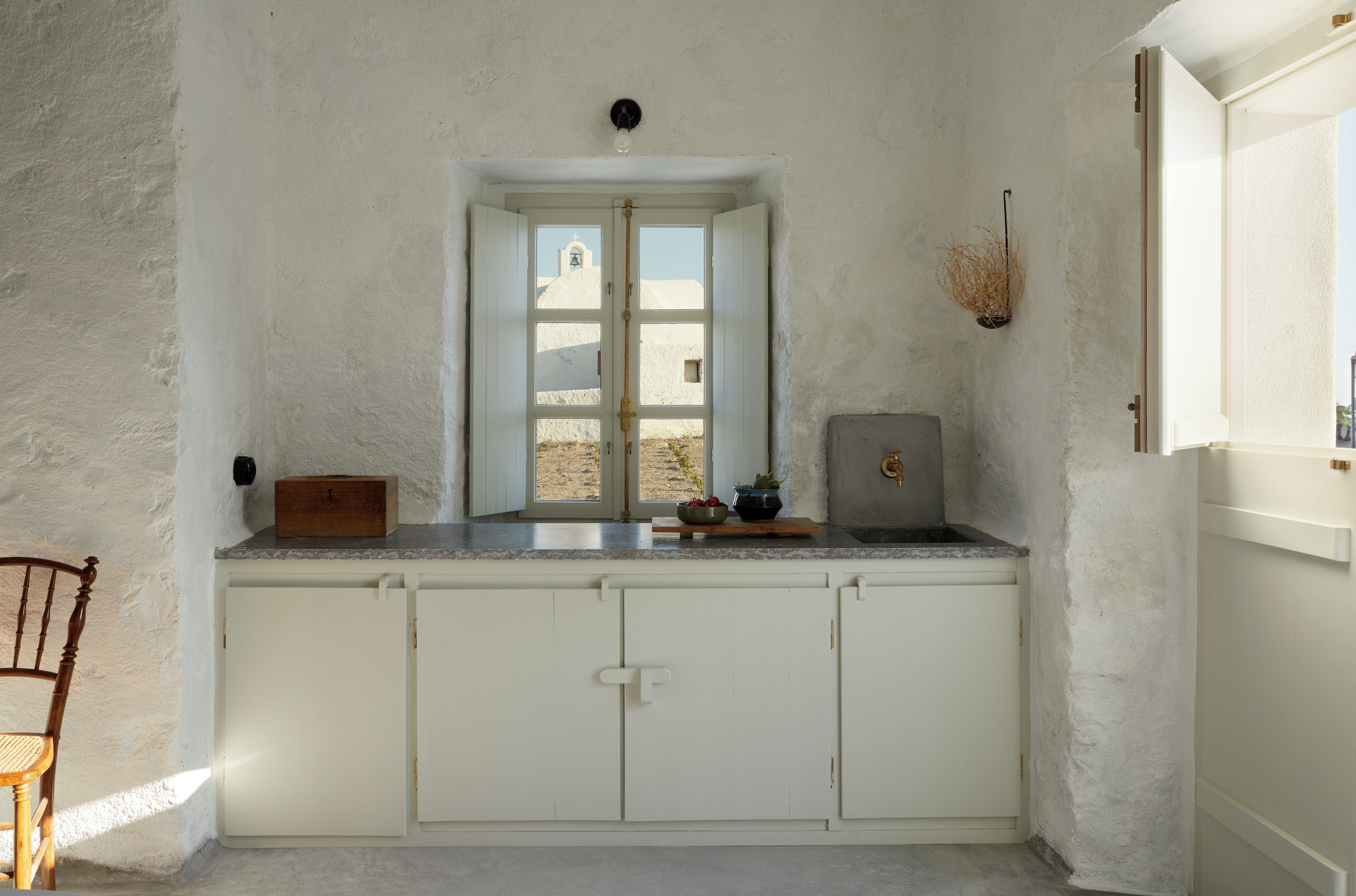 Une cuisine avec des meubles blancs, une chaise en bois et une fenêtre donnant sur un bâtiment blanc.