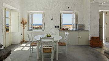 A room with a round table and four chairs, a small kitchen area, and a view of the sea through open windows.