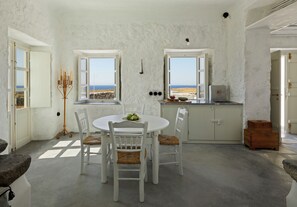 A room with a round table and four chairs, a small kitchen area, and a view of the sea through open windows. - Santa Irini Retreat (Therasia)