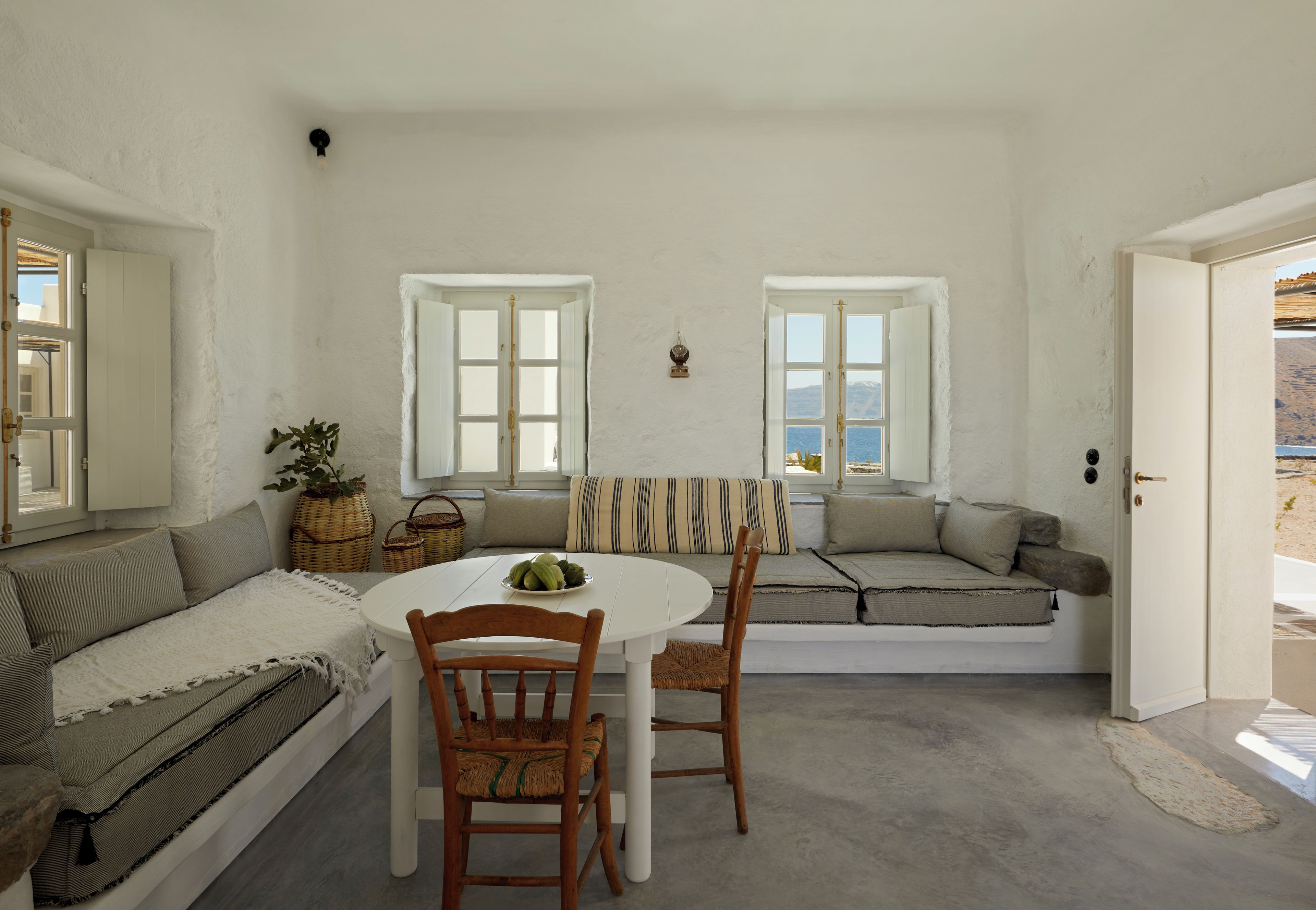 A room with a white interior, a round table, two wooden chairs, a sofa, and a window with a view of the sea.