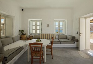 A room with a white interior, a round table, two wooden chairs, a sofa, and a window with a view of the sea. - Santa Irini Retreat (Therasia)