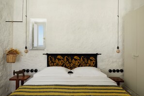 A bedroom with a bed, a window, a hanging light fixture, and a wall-mounted power outlet. - Santa Irini Retreat (Therasia)