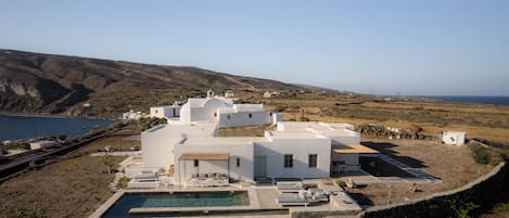 A white villa with a swimming pool, surrounded by a stone wall, overlooking a coastal landscape.