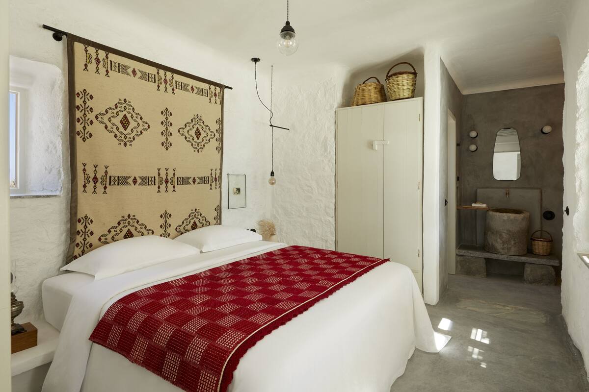 A bedroom with a large bed, a wardrobe, a mirror, and a small bathroom with a sink and a basket.