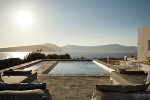A pool with a view of the sea and mountains, surrounded by lounge chairs and a modern building.