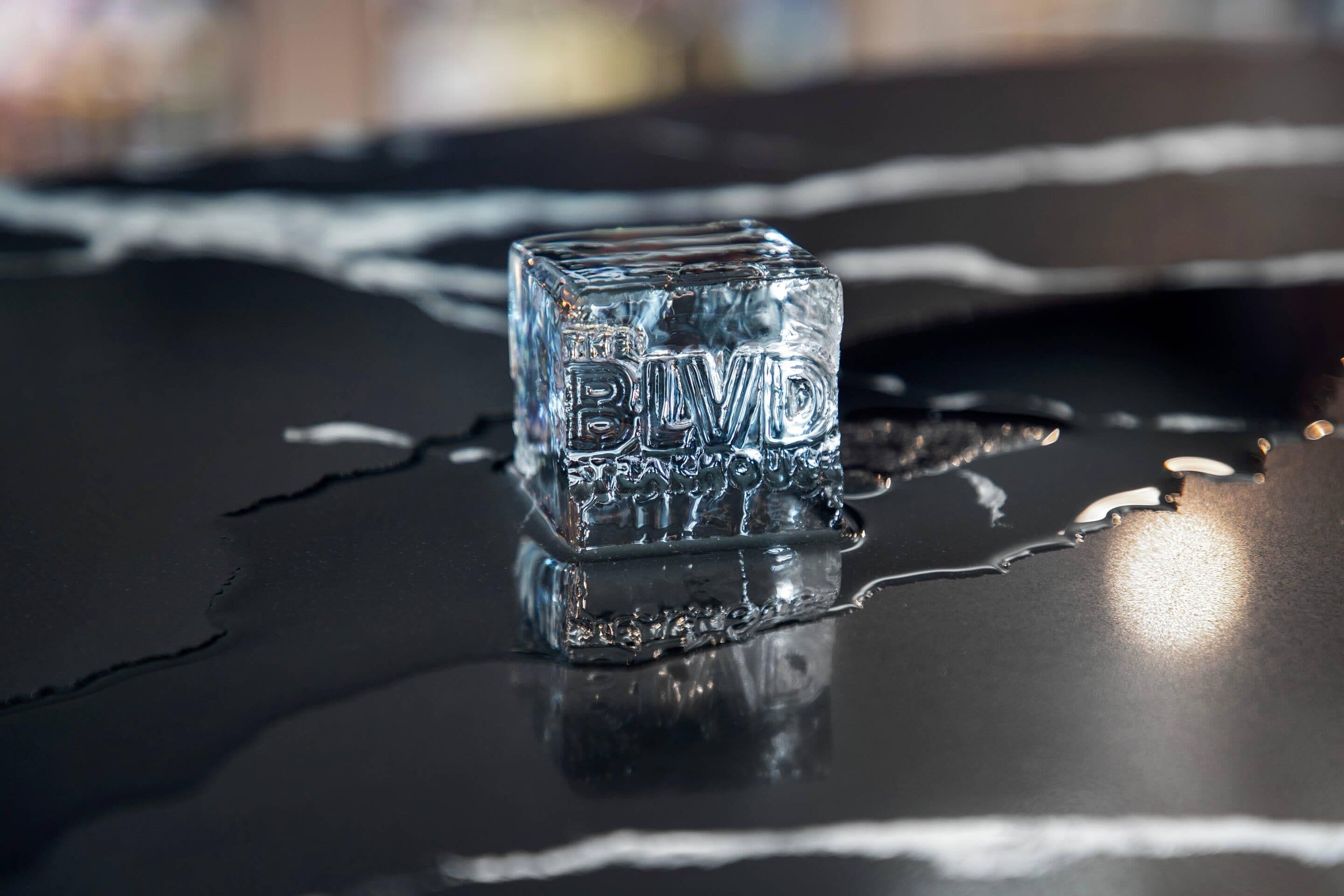 A clear ice cube with the text "THE BLDG" on it, placed on a reflective surface with water droplets around it.