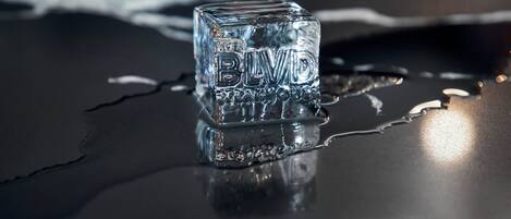 A clear ice cube with the text "THE BLDG" on it, placed on a reflective surface with water droplets around it.