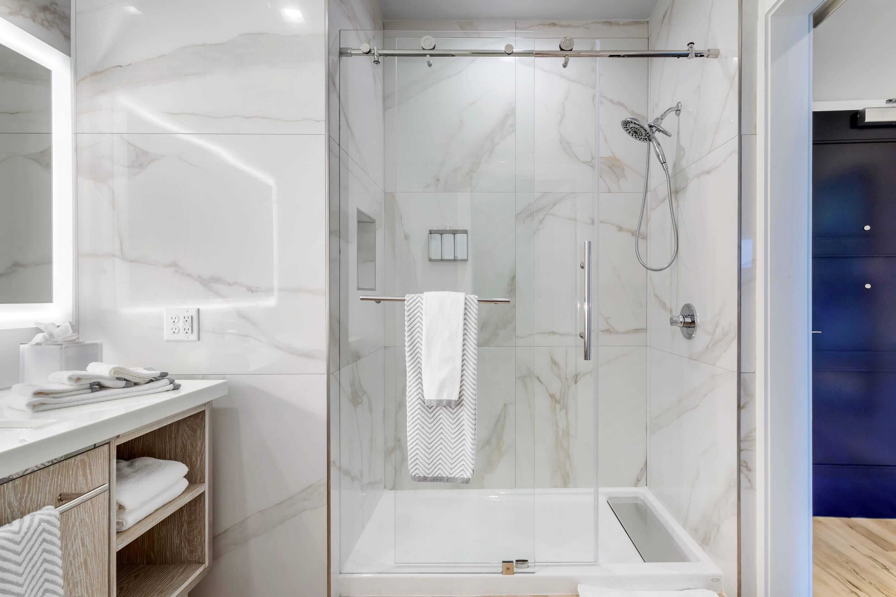 A modern bathroom with a glass-enclosed shower, a white marble wall, a wooden vanity, and a towel rack.