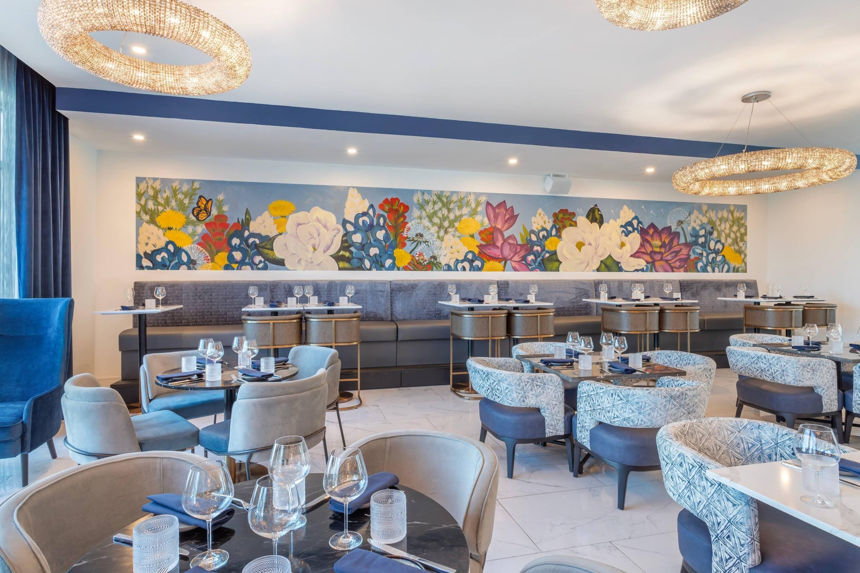 Un restaurante moderno con mesas redondas, sillas azules con diseño y un gran mural floral en la pared.