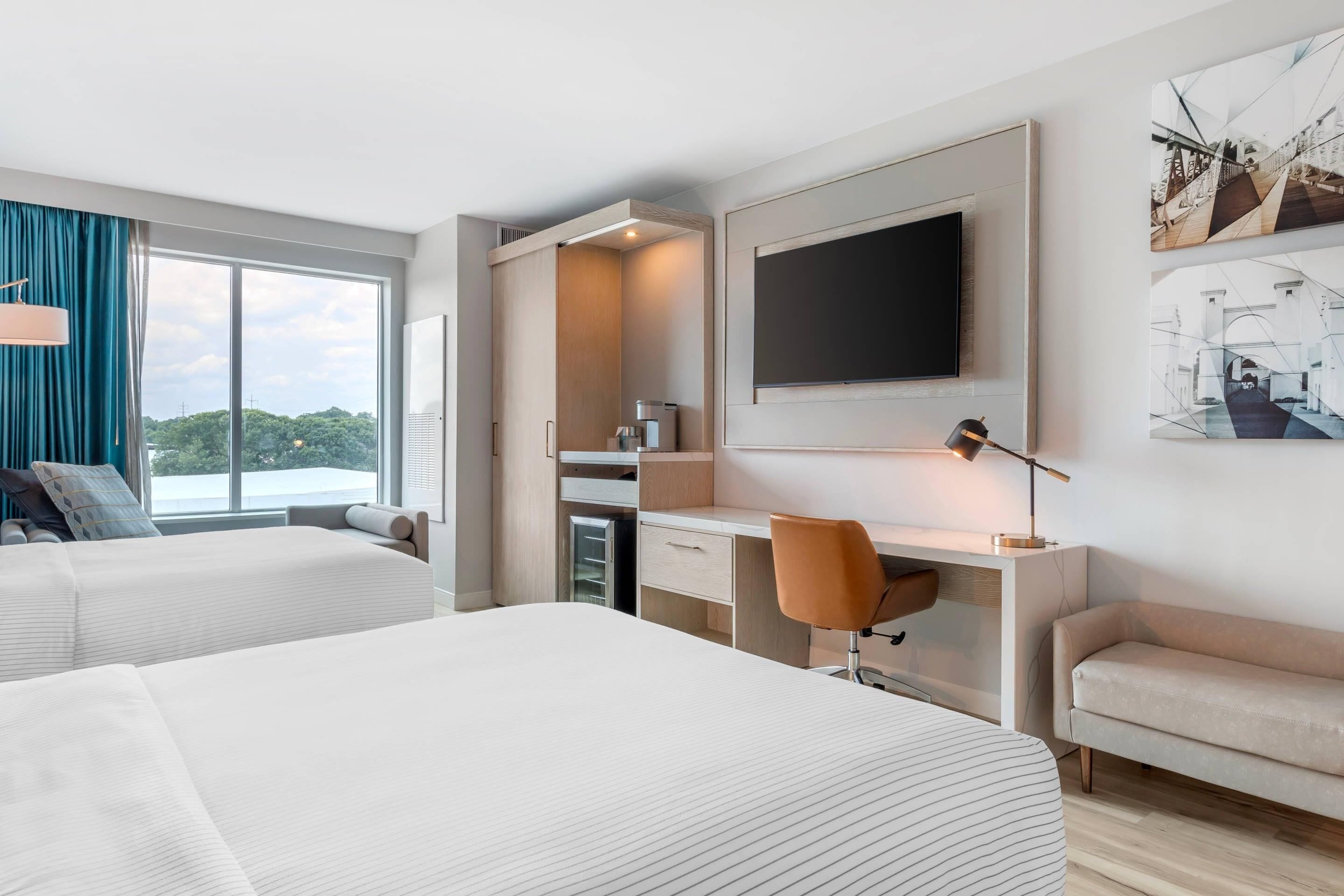 A modern hotel room with two beds, a desk, a chair, a TV, and a large window offering a city view.
