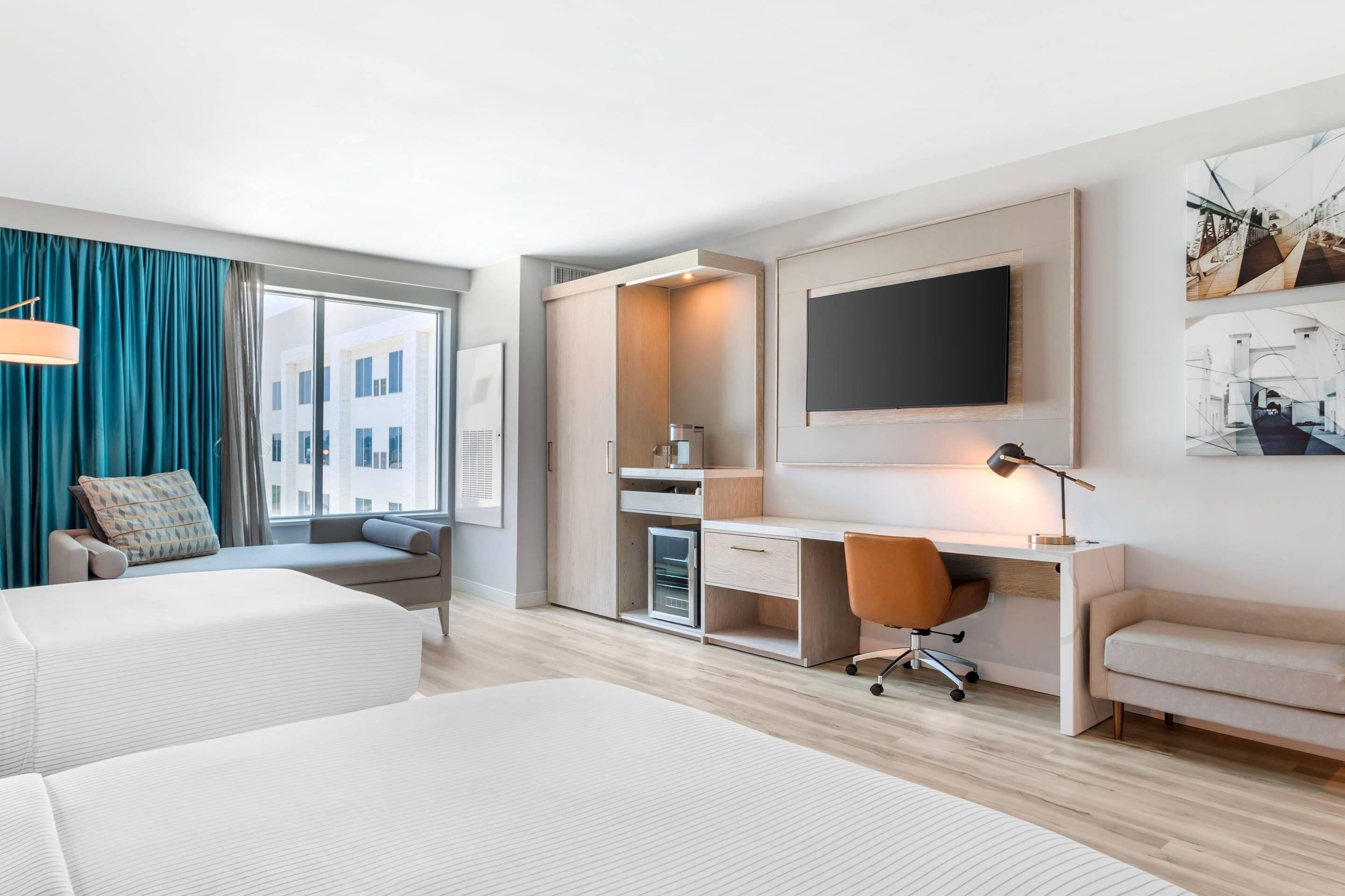 A modern hotel room with two beds, a desk, a chair, a TV, and a large window with curtains.