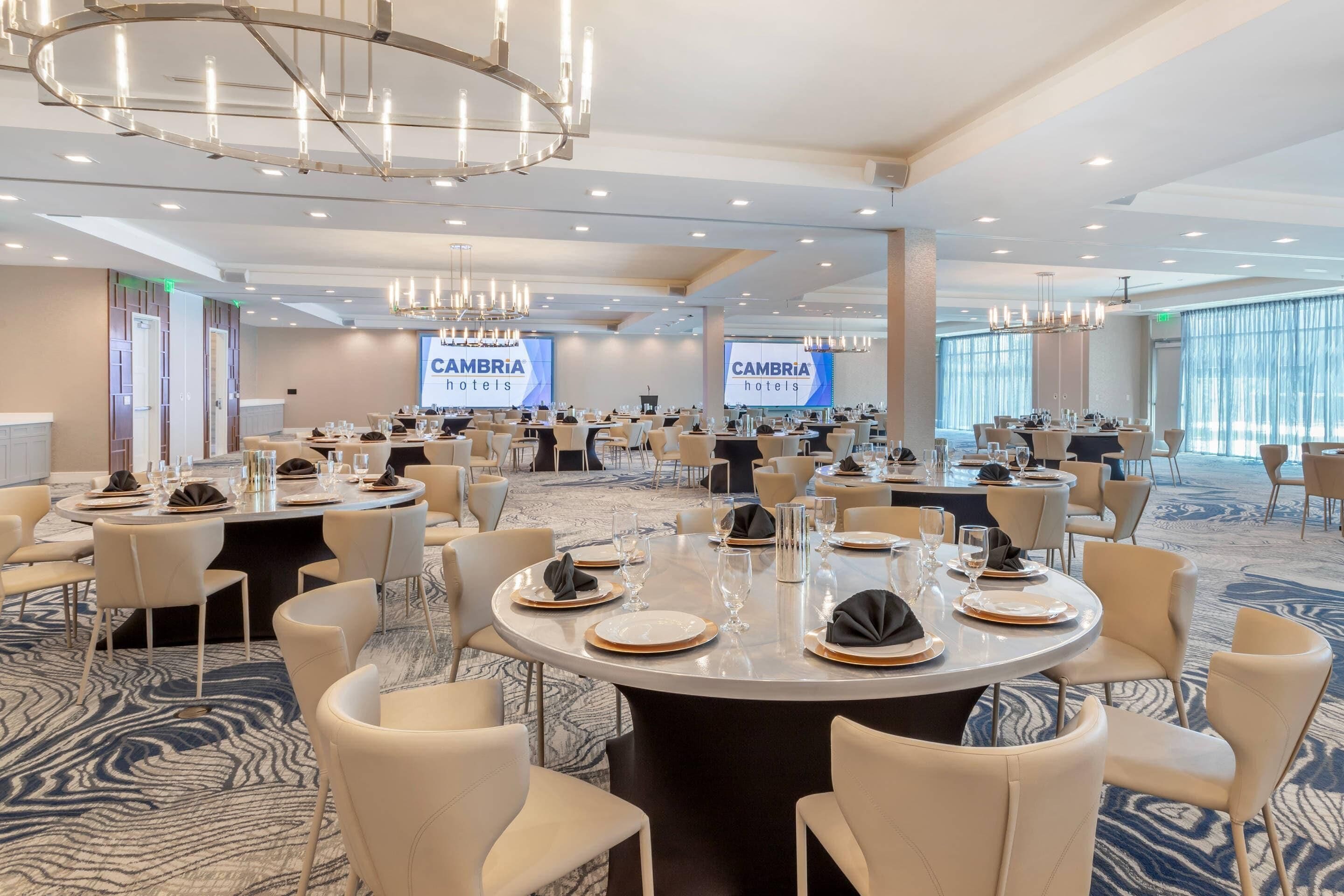 Une salle de conférence équipée de tables rondes, de chaises et d’un grand écran affichant le nom « CAMBRIA hotels ».