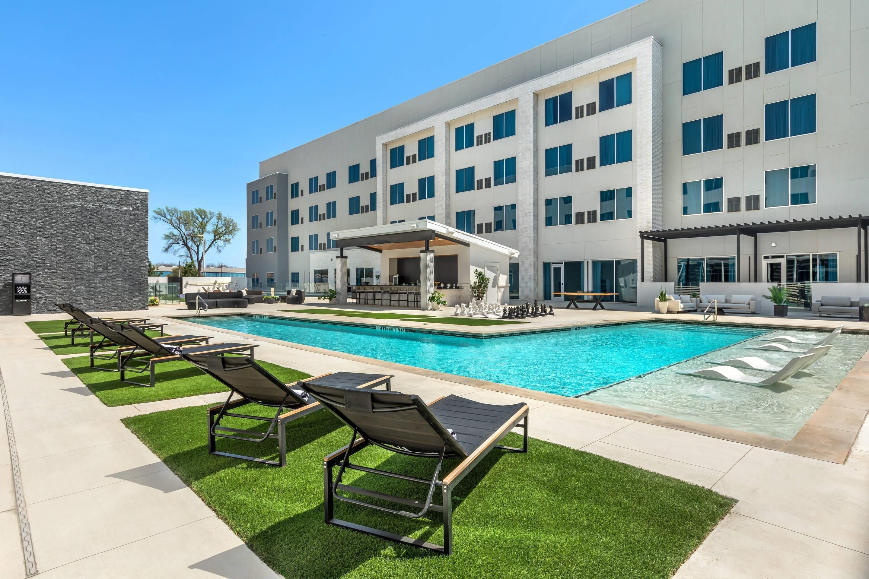 A modern building with a swimming pool, outdoor lounge chairs, and a covered seating area.