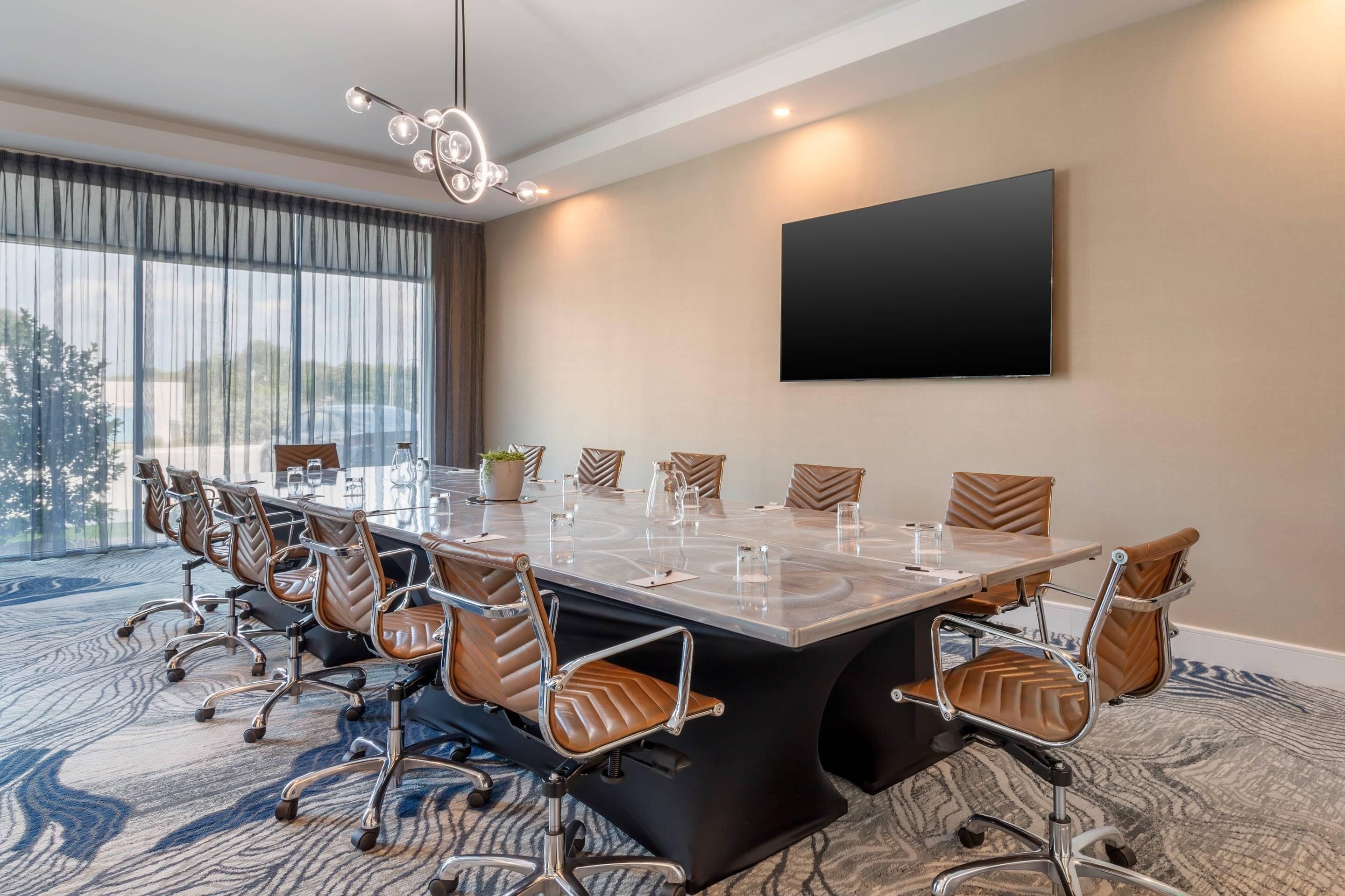 A conference room with a long table, brown leather chairs, a large TV on the wall, and a view through the window.