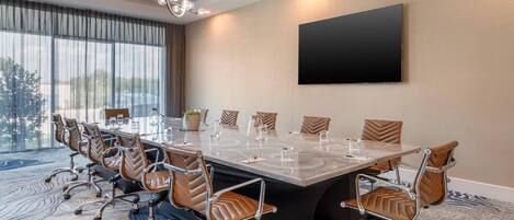 A conference room with a long table, brown leather chairs, a large TV on the wall, and a view through the window.