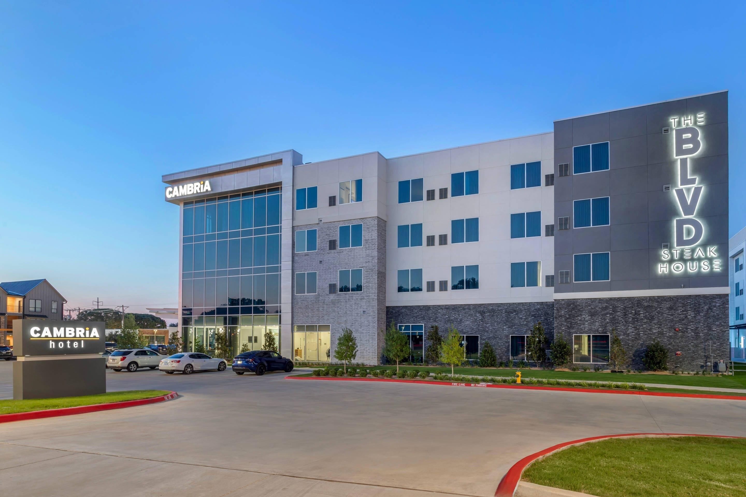 A modern hotel with a glass facade, a sign for 'The BLVD Steak House,' and a parking area with cars.