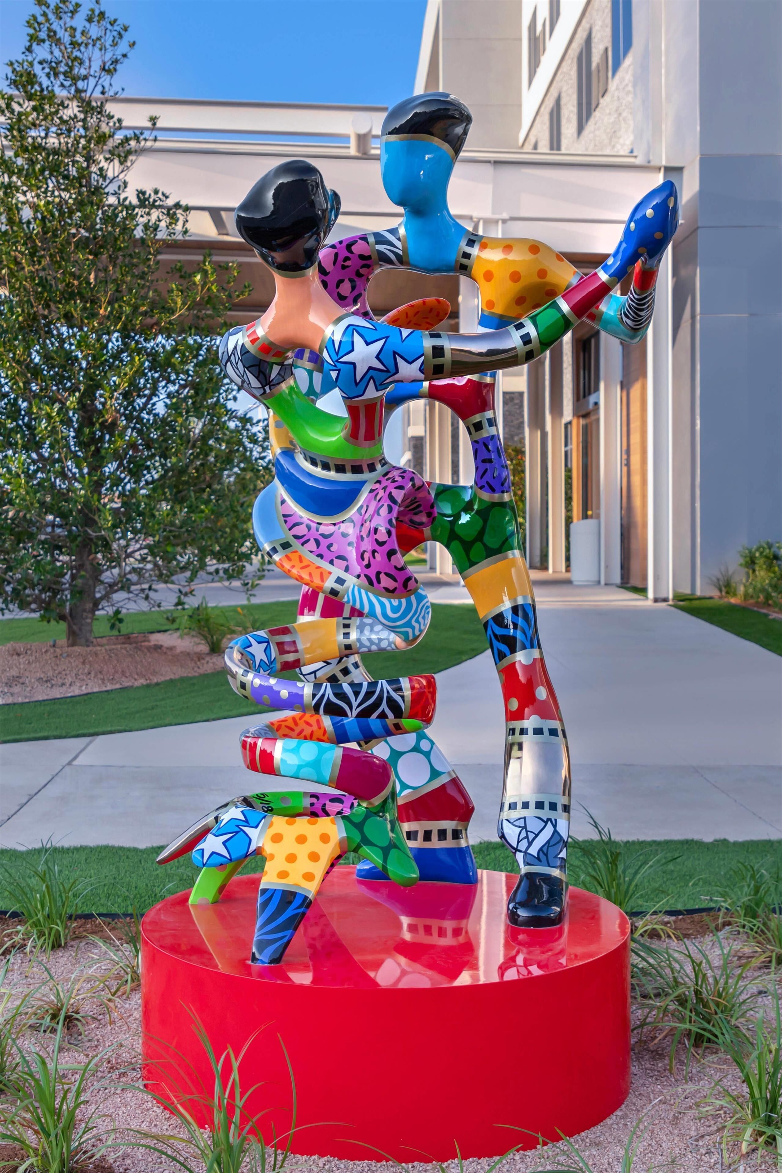 A colorful abstract sculpture in a landscaped area with a building in the background.