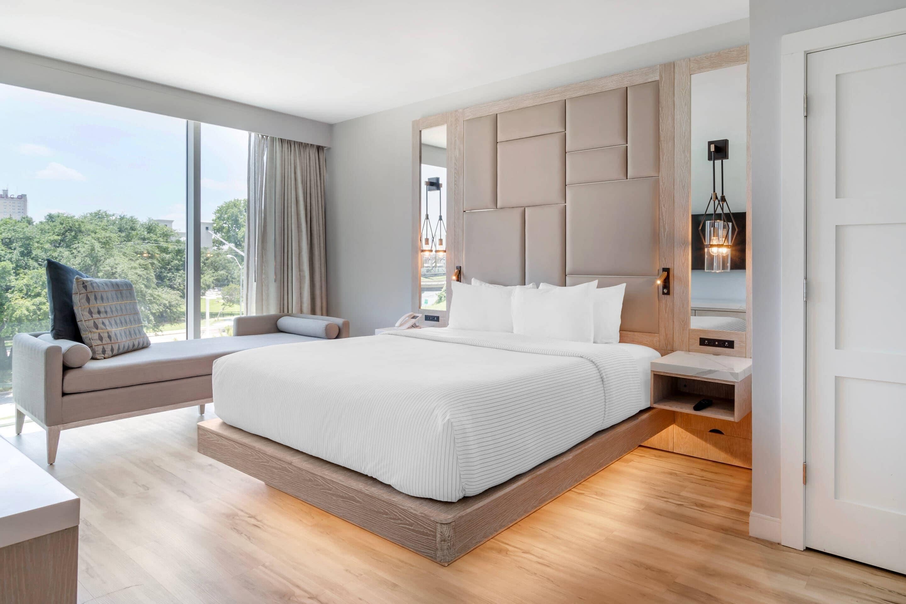 A modern hotel room with a large bed, a seating area with a sofa, a wooden headboard, and a view of the city.