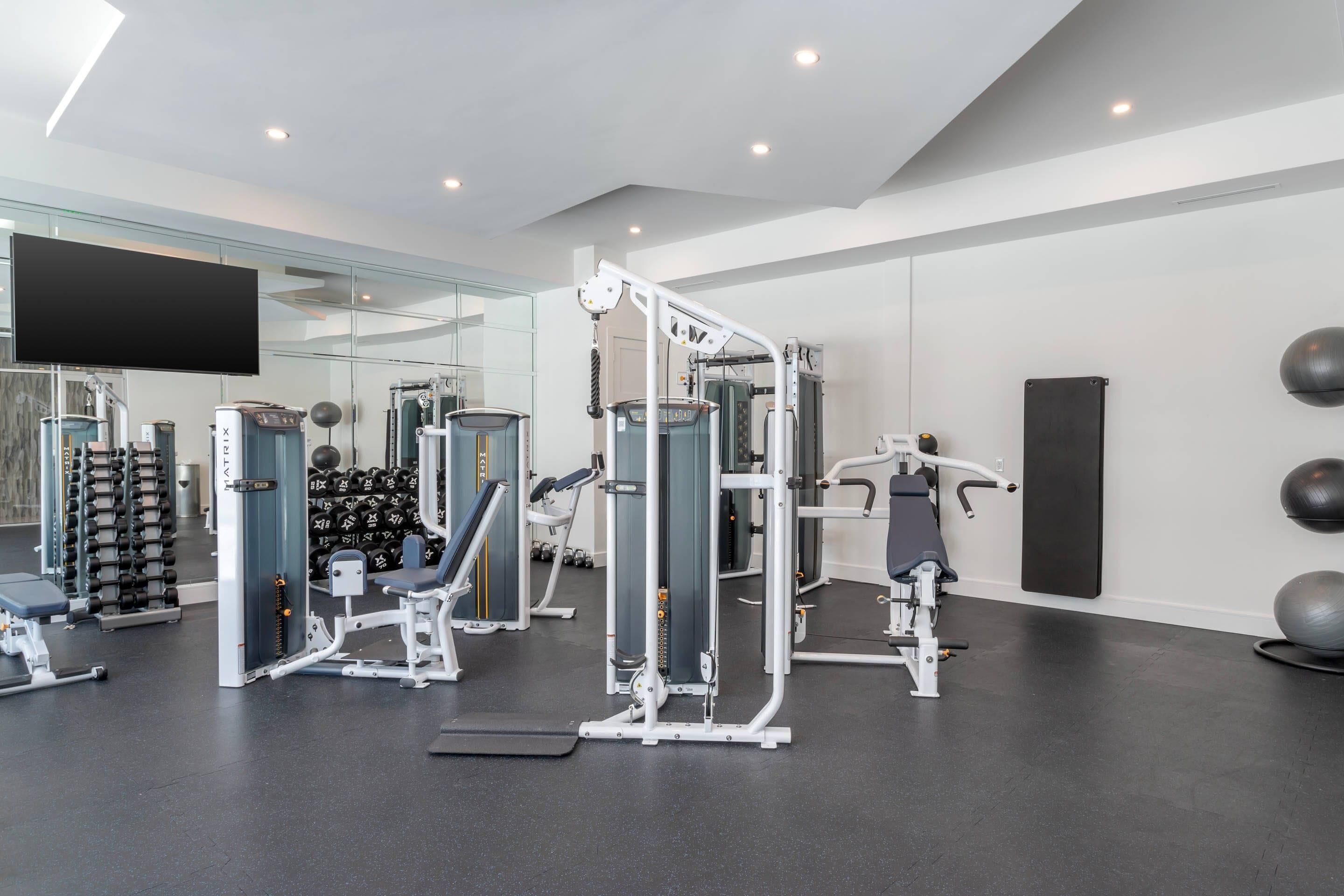 A modern gym with various exercise machines, a large mirror, and a mounted TV.