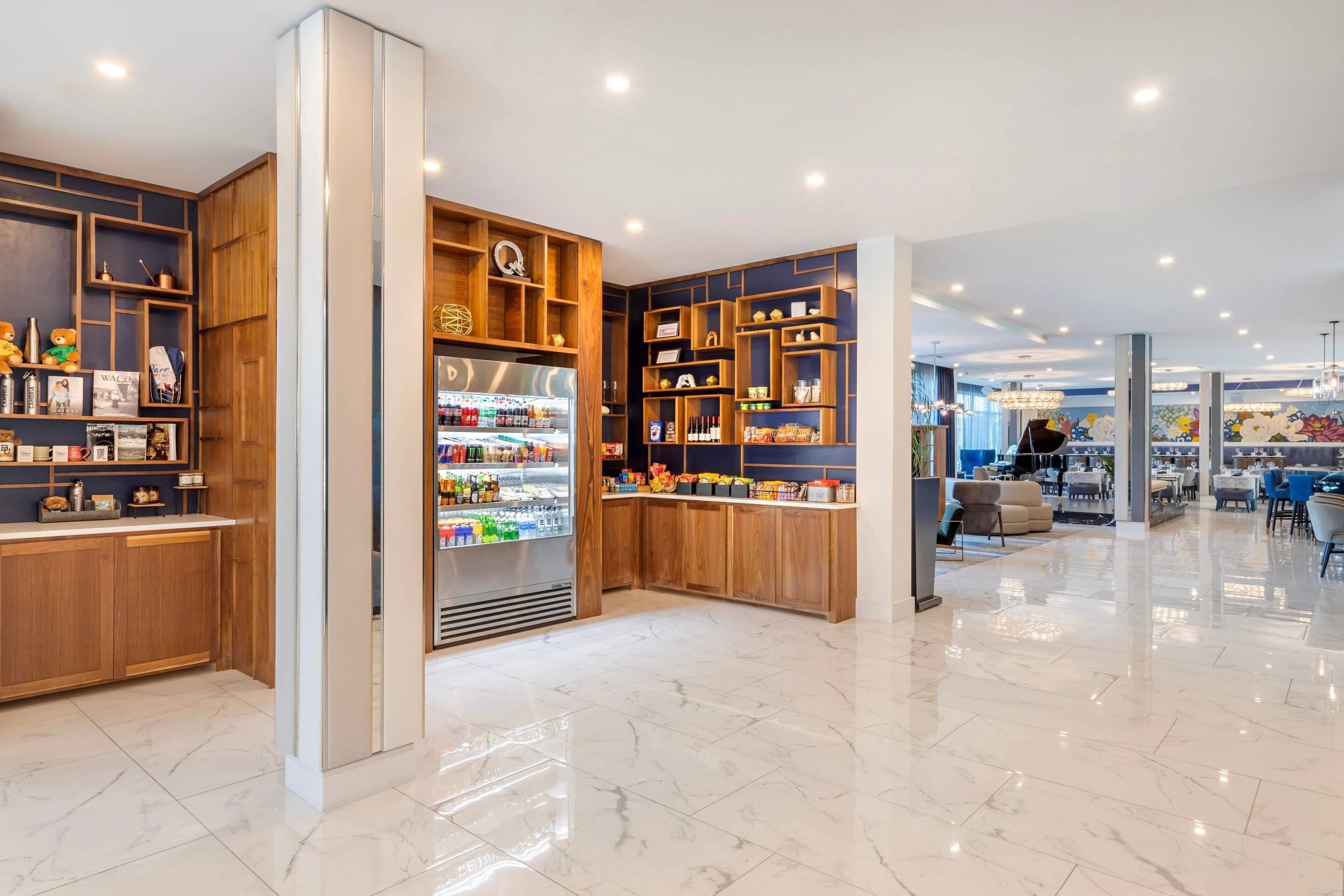 A modern hotel lobby with marble flooring, wooden shelving with various items, a refrigerated display, and a seating area with tables and chairs.