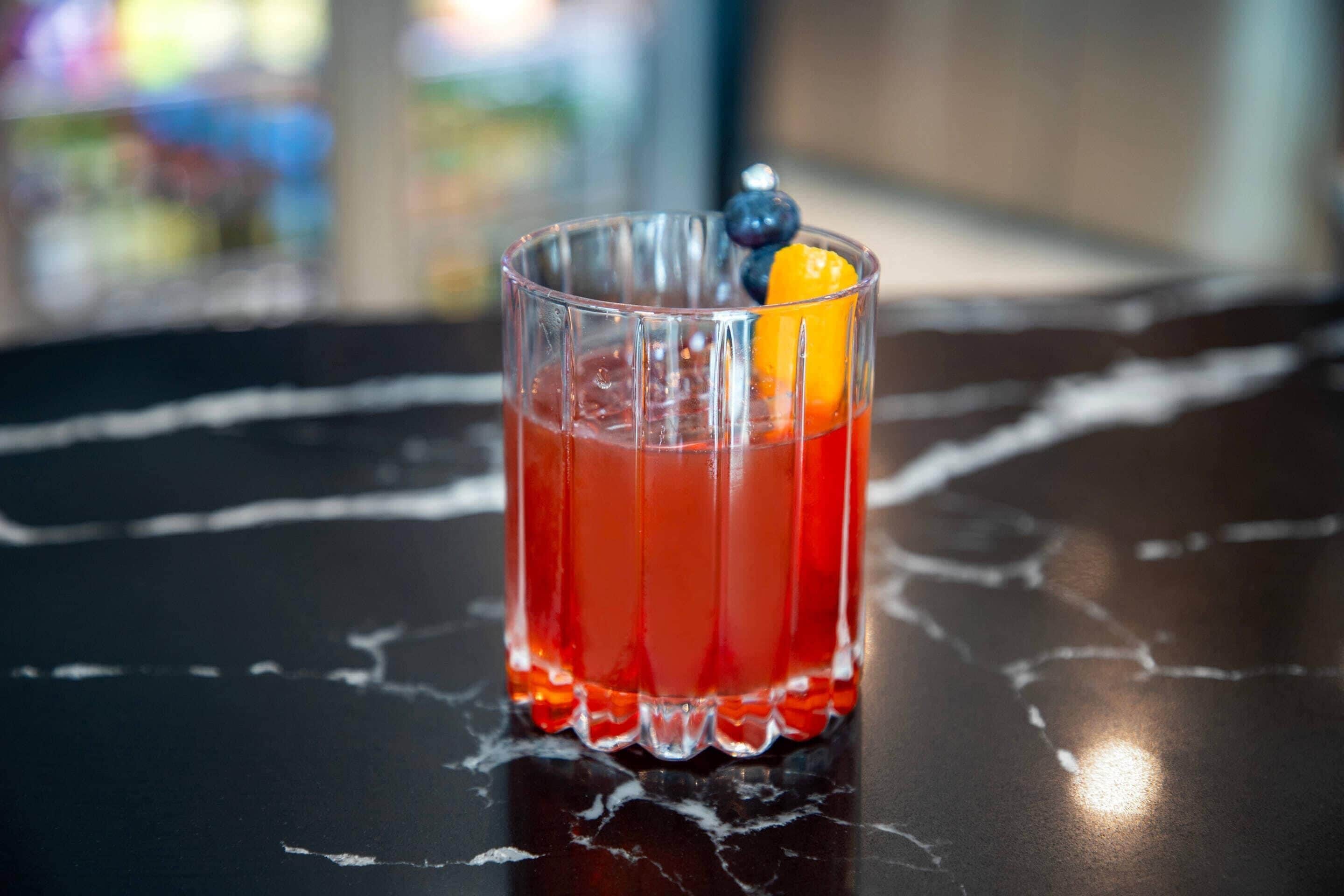 A glass of red beverage with a slice of orange and a blueberry on the rim, placed on a dark surface with visible cracks.