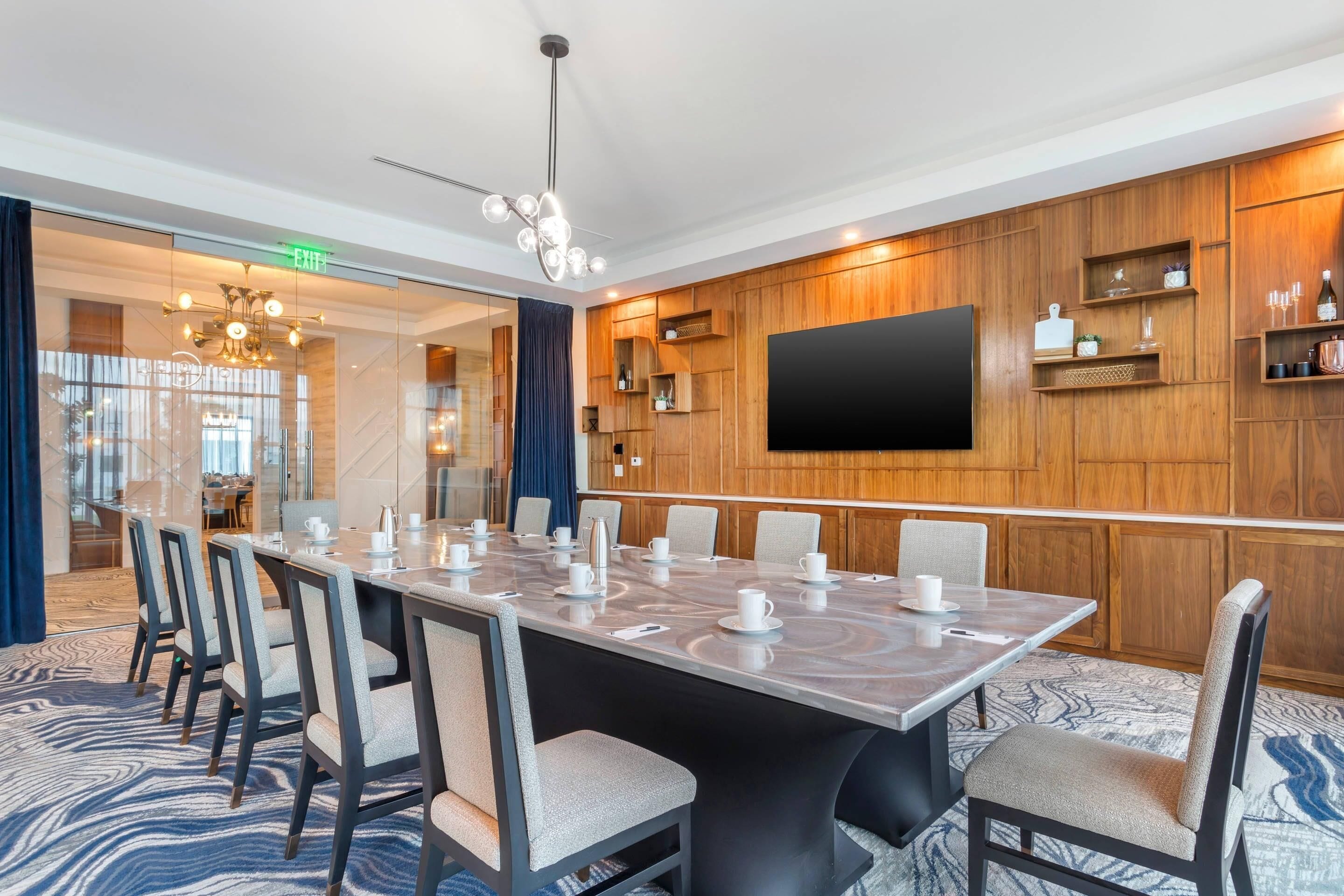 A conference room with a large table, chairs, a flat-screen TV, wooden paneling, and a patterned carpet.