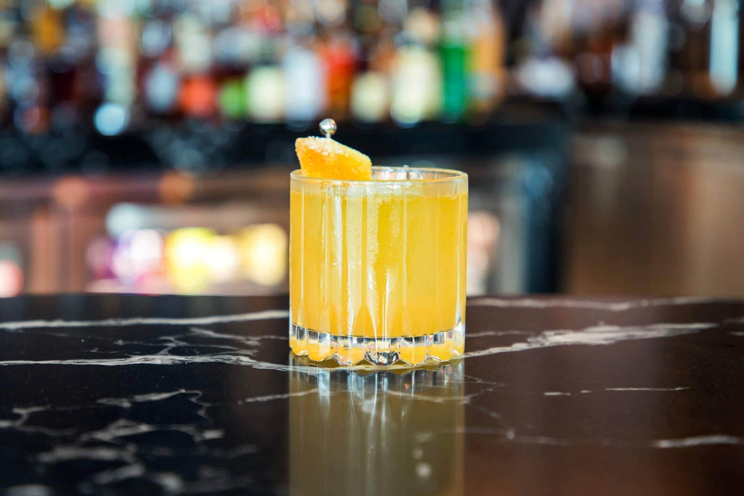 A clear glass of orange juice with a garnish on a reflective surface, likely a bar counter.