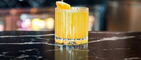 Um copo de suco de laranja, transparente, com um toque decorativo sobre uma superfície refletora, provavelmente um balcão de bar.