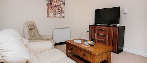 Un salón con un sofá beige, una mesa de centro de madera con un mando a distancia y una taza, un televisor sobre un mueble de madera y un teléfono montado en la pared.