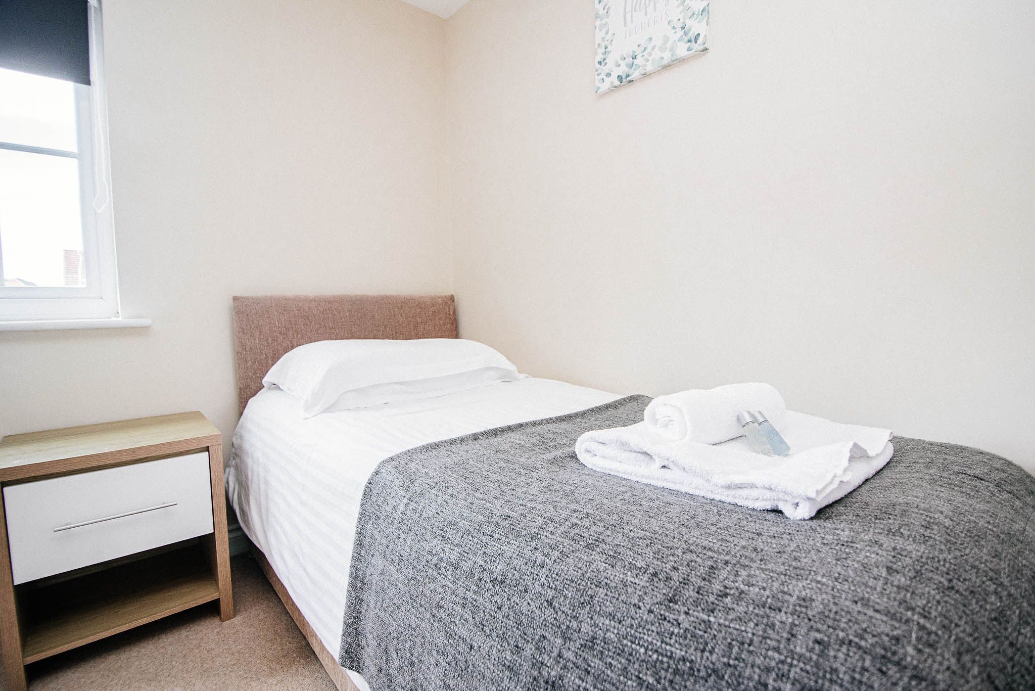A neatly made bed with a grey blanket and white pillows, a wooden nightstand with a drawer, and a window with blinds.