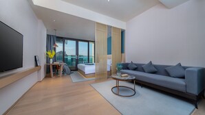 Ein modernes Hotelzimmer mit einem großen Flachbildfernseher, einem grauen Sofa, einem kleinen runden Couchtisch, einem Bett mit Blick und einem großen Fenster.