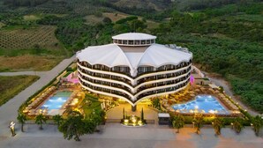 A multi-story hotel with a unique curved roof, multiple balconies, and a central courtyard with pools and outdoor seating. - Seven For Life Thermal Hotel (Kusadasi)