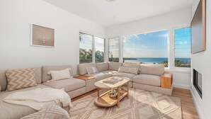 A modern living room with a large sectional sofa, a coffee table, and a view of the sea through large windows.