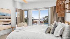A modern hotel room with a large bed, a view of a pool and beach, and a large window.