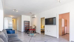 A modern living room with a grey sofa set, a dining area with red chairs, a TV mounted on the wall, and a bookshelf.
