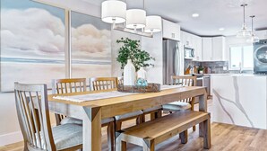 A modern kitchen with white cabinets, a wooden dining table, and pendant lights. - Elegant Coastal-Chic Condo: Pool, Gorgeous Views & Steps to the Pier (Cocoa Beach)