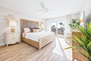 A bedroom with a wooden bed, a nightstand, a desk, and a balcony with a view of palm trees.