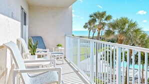 A balcony with white chairs and a small table, overlooking a pool and palm trees. - Elegant Coastal-Chic Condo: Pool, Gorgeous Views & Steps to the Pier (Cocoa Beach)