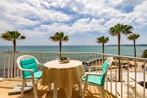 Un balcón con mesa y sillas, con vista a una playa con palmeras y el mar.